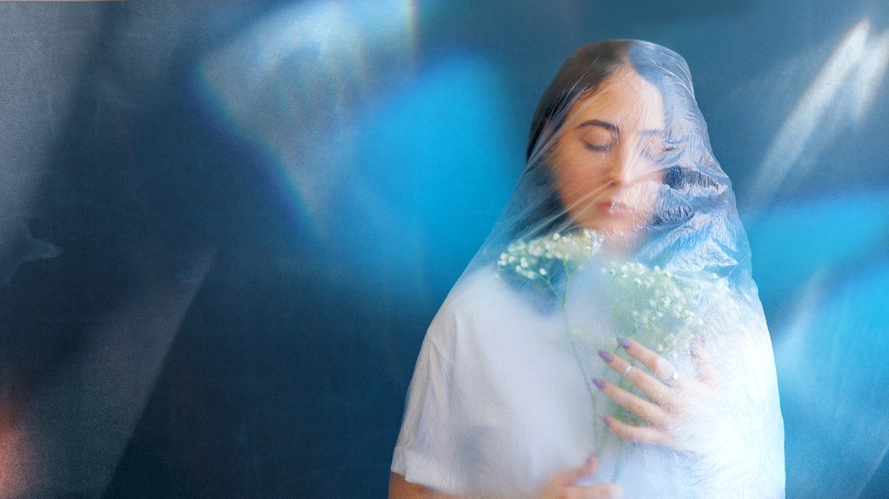 Eine Frau mit Blumen in der Hand, die eine Plastikfolie als Schleier trägt. | Bildquelle: © BR/UFA Documentary/Shutterstock/silverkblackstock